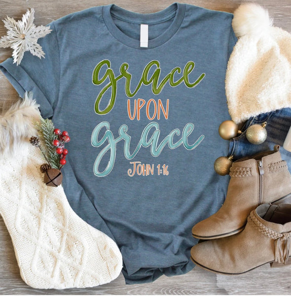 Flat lay photo of our grace upon grace tee. The tee is light blue with lettering on it of green, orange, and bright blue. The flat lay photo is decorated with snowflakes, beige booties, a white stocking, gold ornaments, and light wash jeans.
