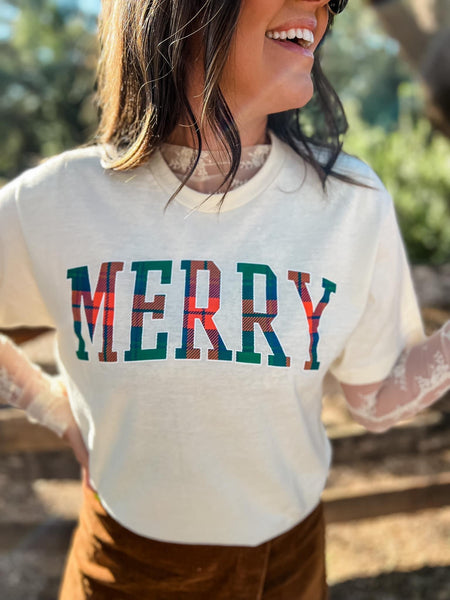 Model wearing our ivory merry plaid tee. The tee is ivory colored and short sleeved. MERRY is written across the chest in bold filled in with blue, orange green plaid. Model paired it with a lace turtleneck underneath and a brown corduroy skirt.