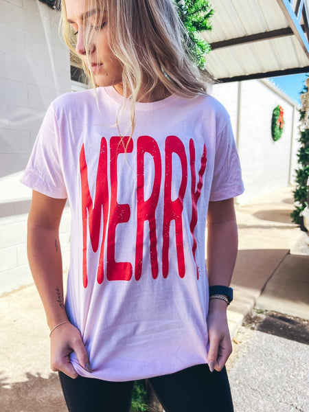 Model wearing our Pink Merry Tee. Tee is short sleeve, light pink, and has the word MERRY in red largely written across it.