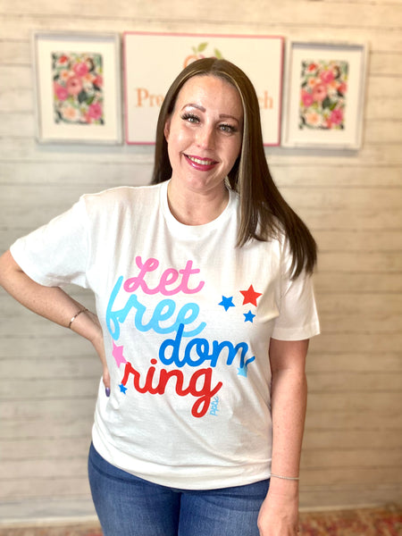 Model wearing our Let Freedom Ring Tee. The tee is short sleeved and has cursive "let freedom ring" written on it. There is stars around the words. It consists of colors pink, aqua, blue, and red. The tee is white.