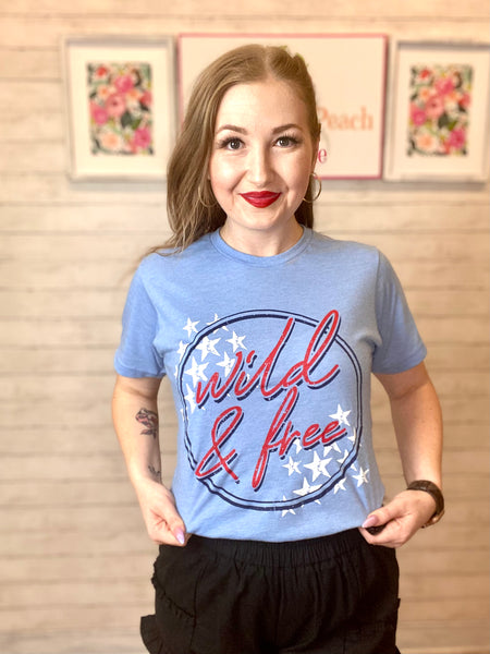 Model wearing our Blue Wild & Free Tee. The top is short sleeve and has a scoop neckline. The top has a dark blue circle with wild & free written in red cursive and white stars on the top left and bottom right corners.