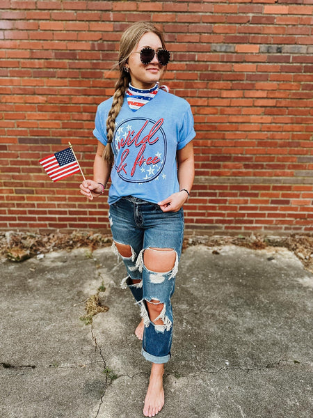Model wearing our Blue Wild & Free Tee. The top is short sleeve and has a scoop neckline. The top has a dark blue circle with wild & free written in red cursive and white stars on the top left and bottom right corners.  