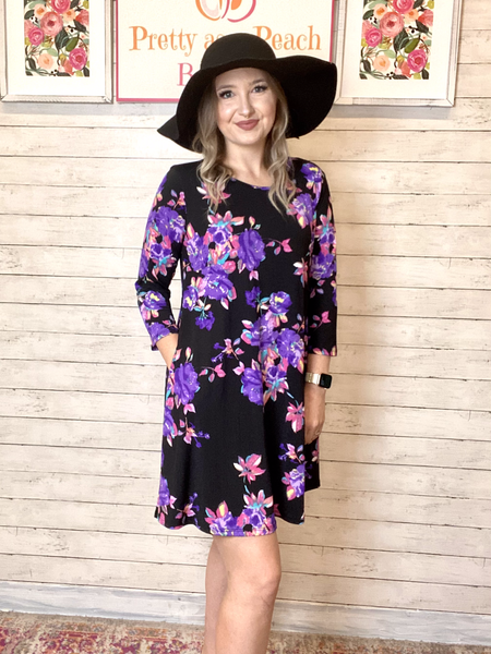 Model wearing our Black & Purple Floral Print Pocket Dress. The dress is 3/4 sleeve length and is above knee length. The dress is black with purple, pink, and blue blossoms. The dress also has pockets. Model pairs the dress with a black hat.