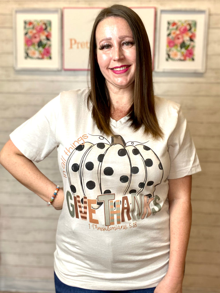 Model wearing our tee that says "in all things, give thanks. 1 Thessalonians 5:28". The tee is short sleeve and v neck with a pumpkin with polka dots. Each letter of Give thanks has patterns.