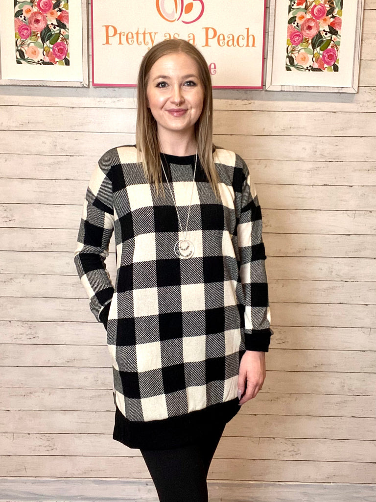 Model wearing our Black & Ivory Buffalo Plaid Tunic Sweater. The sweater is long sleeved, with pockets, and legging friendly. Model pairs the tunic sweater with black leggings and a long silver necklace.