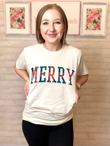 Model wearing our ivory merry plaid tee. The tee is ivory colored and short sleeved. MERRY is written across the chest in bold filled in with blue, orange green plaid.