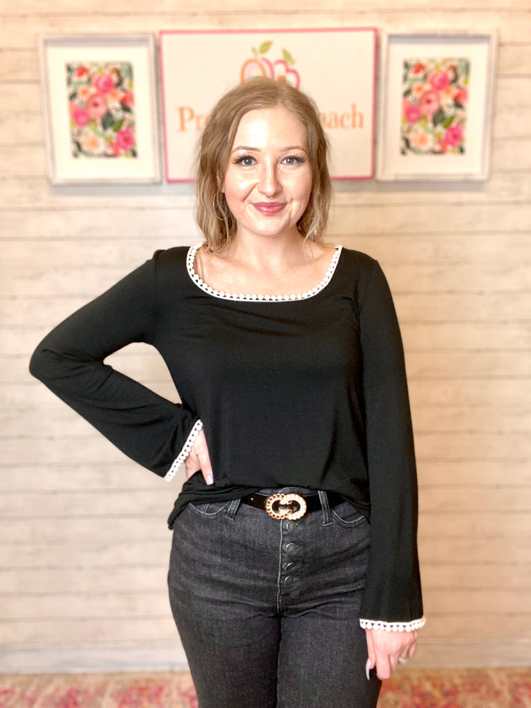 Model wearing a black crochet accent top. Top is long sleeve and black with white crochet accent on the neckline and wrist. Model paired the top with high waisted black jeans.