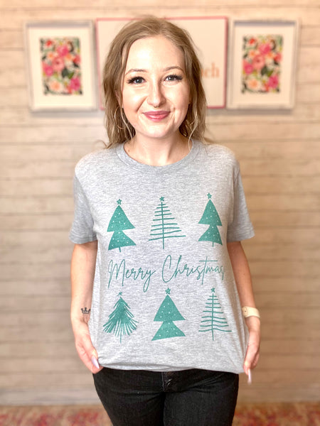 Model wearing our Merry Christmas Tree Tee HOLIDAY FINAL SALE. Tee is gray with green trees and Merry Christmas written across the middle. Tee is short sleeve.