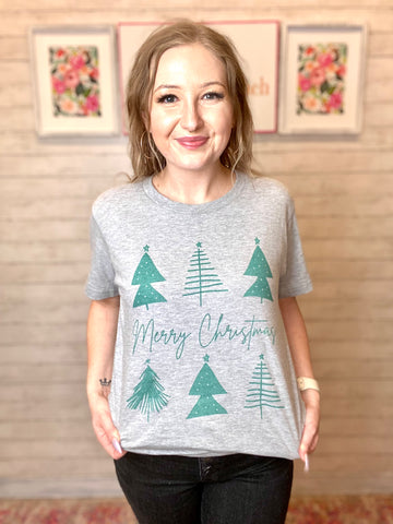 Model wearing our Merry Christmas Tree Tee HOLIDAY FINAL SALE. Tee is gray with green trees and Merry Christmas written across the middle. Tee is short sleeve.