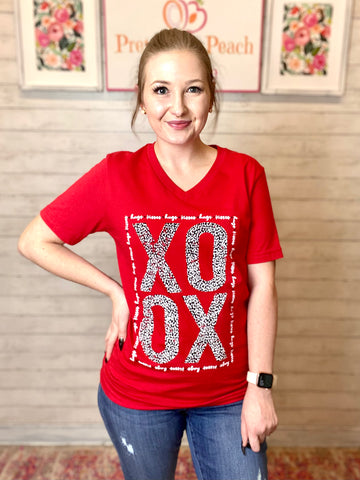 Model wearing our red xoxo tee. The tee is short sleeve and bright red with v-neck. The letters xoxo are in bold with black and white leopard print making them. hugs and kisses is written in cursive around the bold letters making a rectangle border.