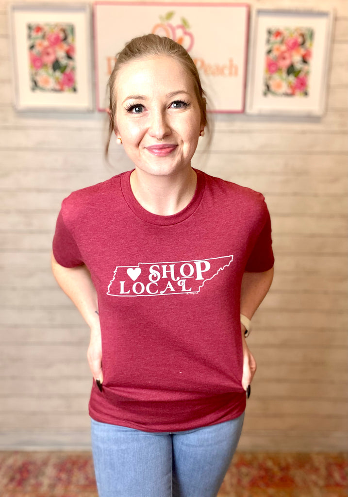 Model wearing our cranberry shop local TN tee. Tee is short sleeve and has the outline of the TN state in white with "Shop local" written in funky lettering with a white heart to the left.