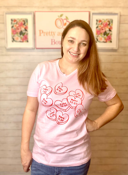 Model wearing our Pink Faith Conversation Hearts Tee. The tee is short sleeve with conversation hearts all over it. Inside each heart is a Bible verse number and chapter. The model pairs the top with light wash denim jeans.