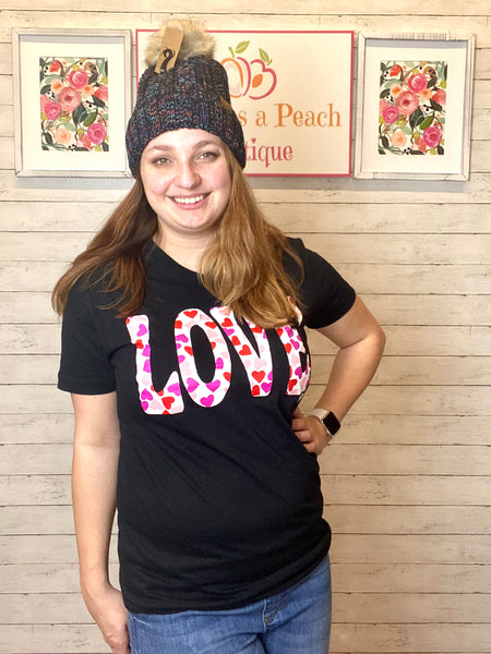 Model wearing our love heart tee. The tee is short sleeve and black with LOVE written in bold white with red and pink hearts inside. The tee is a subtle v-neck and can be paired with so many things. Our model pairs the top with jeans and a pom pom beanie.