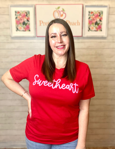 Model wearing our red sweetheart tee. The tee is short sleeve with a normal high scoop neck. The tee is red with pink cursive lettering spelling sweetheart. 
