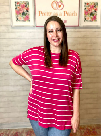 Model wearing our Fuchsia Oversized Striped Tunic. The top is elbow length and oversized. The top is fuchsia with white stripes horizontally. Model pairs the top with light wash denim jeans.