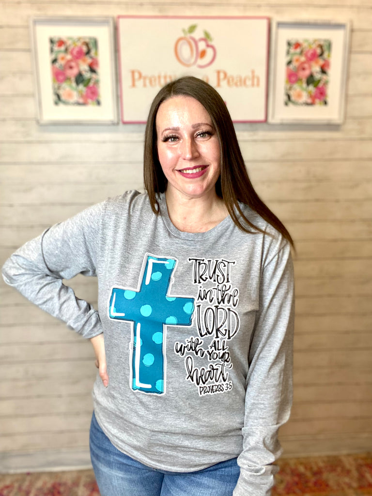Model wearing our Trust in the Lord Long Sleeve Tee. The tee is gray, long sleeve, and has a subtle scoop neckline. There is cursive lettering on the right side and a bolded cross on the left filled in blue with light blue polka dots.