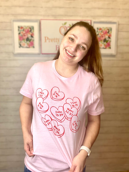 Model wearing our Pink Faith Conversation Hearts Tee. The tee is short sleeve with conversation hearts all over it. Inside each heart is a Bible verse number and chapter. The model pairs the top with light wash denim jeans.