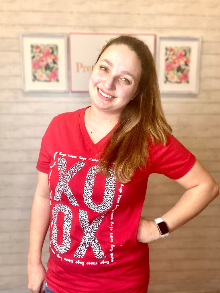 Model wearing our red xoxo tee. The tee is short sleeve and bright red with v-neck. The letters xoxo are in bold with black and white leopard print making them. hugs and kisses is written in cursive around the bold letters making a rectangle border.