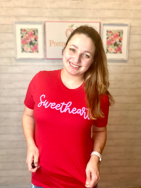 Model wearing our red sweetheart tee. The tee is short sleeve with a normal high scoop neck. The tee is red with pink cursive lettering spelling sweetheart.