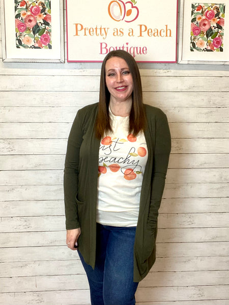 Model wearing the olive 3/4 sleeve ruffle cardigan. Cardigan is loose and soft with a ruffle hem and pockets. Cardigan gives a fun and girly look. Model pairs the cardigan with a graphic tee and dark denim.