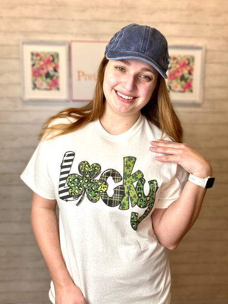 Model wearing our lucky print tee. The tee is short sleeve and white with the word lucky. The word is bold and filled in with different green prints inside. The "u" is a clover.