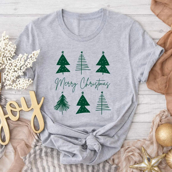 Flat lay photo of our Merry Christmas Tree Tee. Tee is gray with green trees and Merry Christmas written across the middle. Tee is short sleeve. Flat lay also consists of ornaments, burlap, and a joy sign.