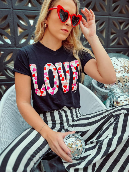 Model wearing our love heart tee. The tee is short sleeve and black with LOVE written in bold white with red and pink hearts inside. The tee is a subtle v-neck and can be paired with so many things.