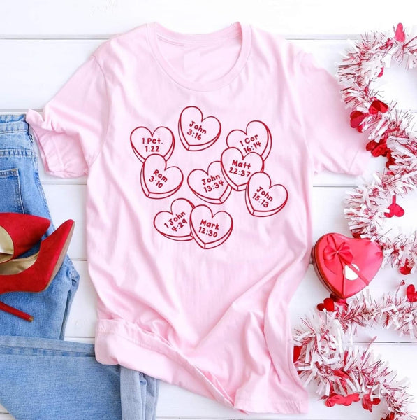 Flat lay photo of our Pink Faith Conversation Hearts Tee. The tee is short sleeve with conversation hearts all over it. Inside each heart is a Bible verse number and chapter. The rest of the flat lay contains red high heels, light denim jeans, a heart garland, and a chocolates heart box.
