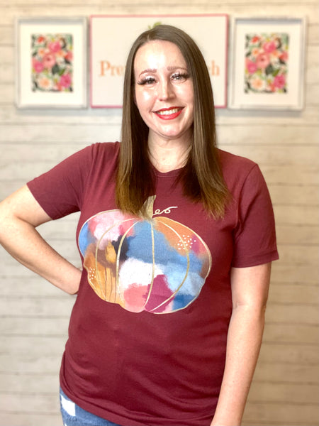 Model wearing our wine multi swatch pumpkin tee. Tee is short sleeve and wine colored with a gold sketch of a pumpkin. Pumpkin is filled in with watercolors of gold, orange, light blue, navy, light pink, fuchsia, white, brown, and black. 