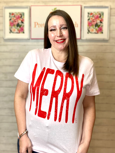 Model wearing our Pink Merry Tee. Tee is short sleeve, light pink, and has the word MERRY in red largely written across it.