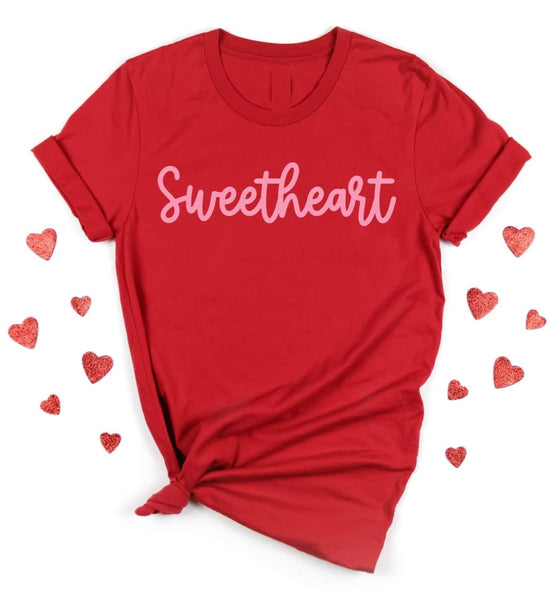 Flat lay photo of our red sweetheart tee. The tee is short sleeve with a normal high scoop neck. The tee is red with pink cursive lettering spelling sweetheart. The flat lay photo has the tee tied in the bottom left corner.
