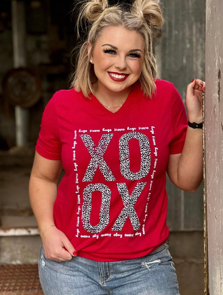Model wearing our red xoxo tee. The tee is short sleeve and bright red with v-neck. The letters xoxo are in bold with black and white leopard print making them. hugs and kisses is written in cursive around the bold letters making a rectangle border.