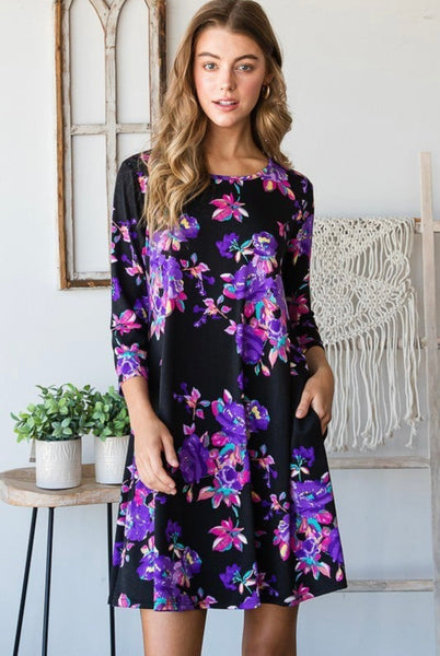 Model wearing our Black & Purple Floral Print Pocket Dress. The dress is 3/4 sleeve length and is above knee length. The dress is black with purple, pink, and blue blossoms. The dress also has pockets. 