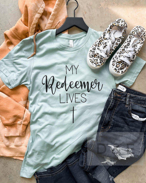 Flat lay photo of our my redeemer lives tee. The tee is light blue and short sleeve with the writing on the chest and a cross at the bottom. 