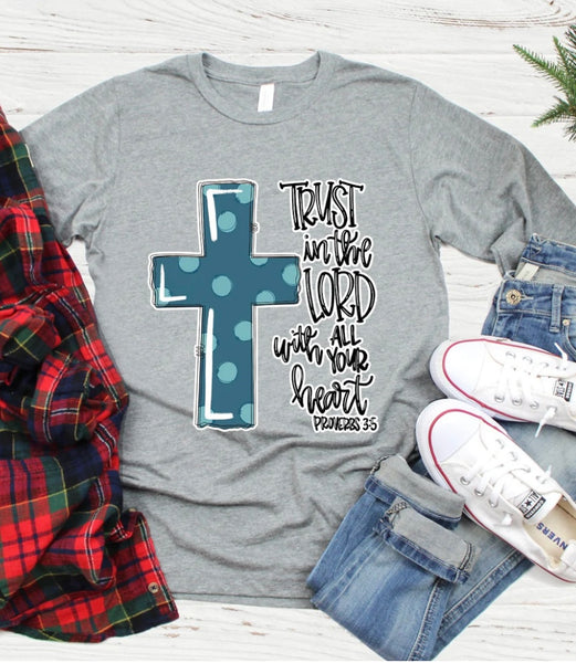 Flat lay photo of our Trust in the Lord Long Sleeve Tee. The tee is gray, long sleeve, and has a subtle scoop neckline. There is cursive lettering on the right side and a bolded cross on the left filled in blue with light blue polka dots.