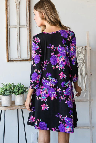 Model wearing our Black & Purple Floral Print Pocket Dress. The dress is 3/4 sleeve length and is above knee length. The dress is black with purple, pink, and blue blossoms. The dress also has pockets.