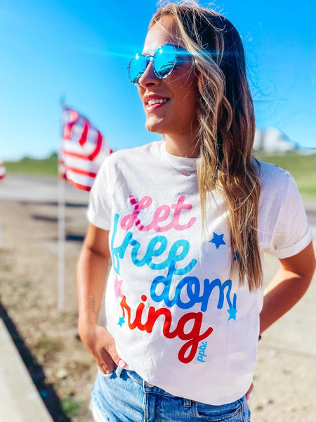 Model wearing our Let Freedom Ring Tee. The tee is short sleeved and has cursive "let freedom ring" written on it. There is stars around the words. It consists of colors pink, aqua, blue, and red. The tee is white.